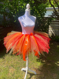 A bright orange and red tulle tutu skirt displayed on a white mannequin outdoors in a backyard setting with grass, trees, and a fence visible.