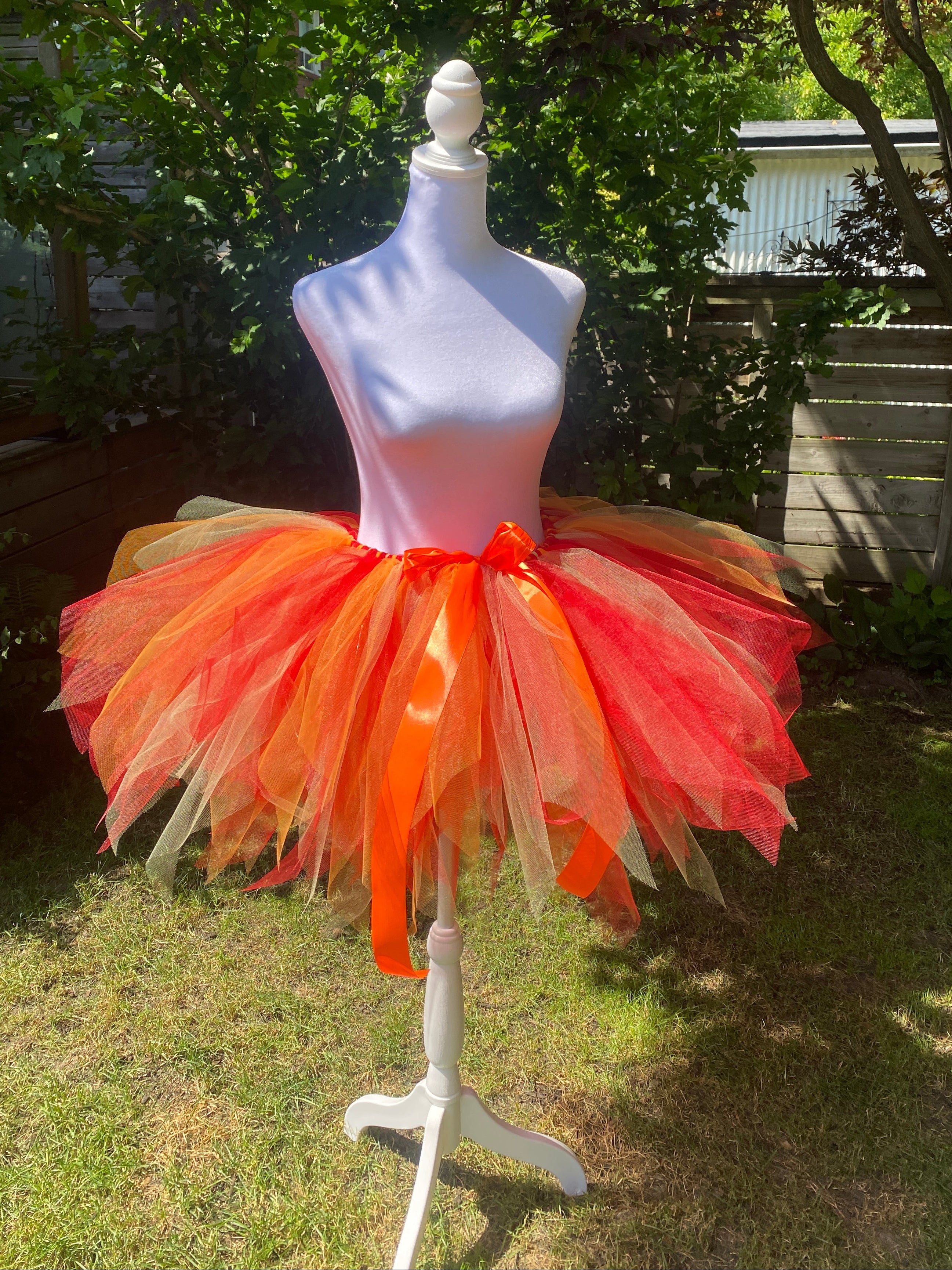 A bright orange and red tulle tutu skirt displayed on a white mannequin outdoors in a backyard setting with grass, trees, and a fence visible.